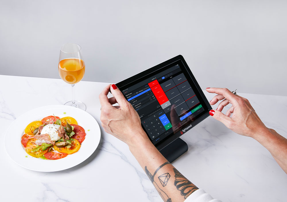 Hände einer Mitarbeiterin die das Restaurant Kassensystem auf einem iPad bedient. Daneben steht ein Getränk sowie ein Teller mit Tomate und Mozzarella